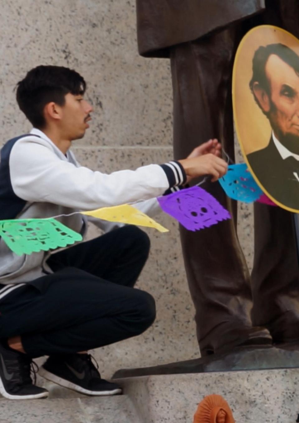 A brown college student ties a string of colorful, celebratory flags in front of a large bronze sculpture of President Lincoln.